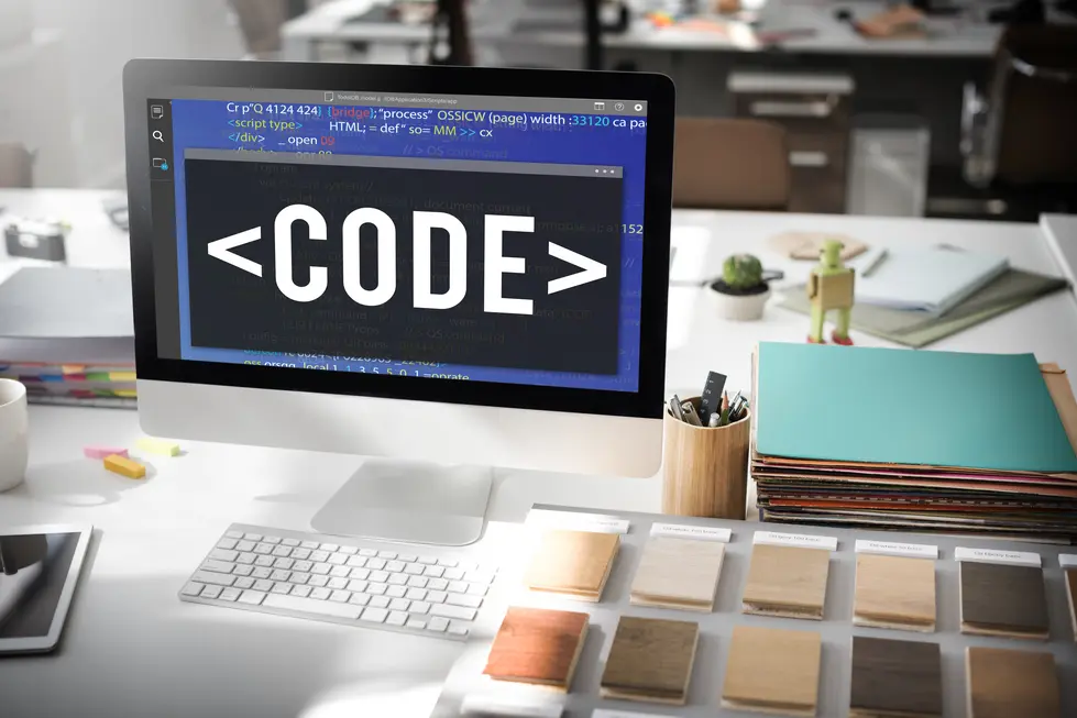 Ein Computerbildschirm mit Code-Editor zeigt den Text "", umgeben von bunten Materialien und Schreibutensilien auf einem Schreibtisch. Der Hintergrund weist wei