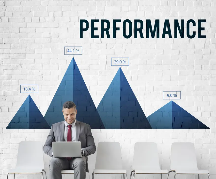 Geschäftsmann sitzt auf einem Stuhl und arbeitet am Laptop, während er vor einer muralen Grafik steht, die verschiedene Leistungskennzahlen mit Prozentangaben zeigt, unter dem Schriftzug "PERFORMANCE". Die Grafik besteht aus stilisierten Bergspitzen in Blau und symbolisiert Wachstum und Performance-Optimierung.