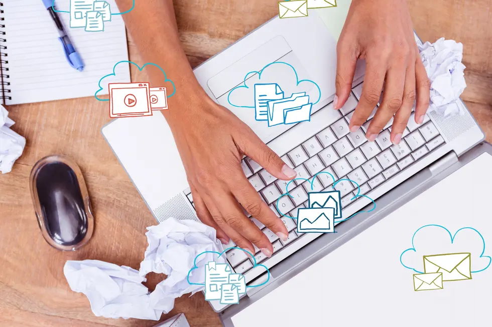 Hände eines Nutzers, die auf einer Laptop-Tastatur tippen, umgeben von zerknülltem Papier und farbigen Grafiken, die verschiedene digitale Inhalte wie Dokumente, Bilder und E-Mails symbolisieren, auf einem Holztisch.