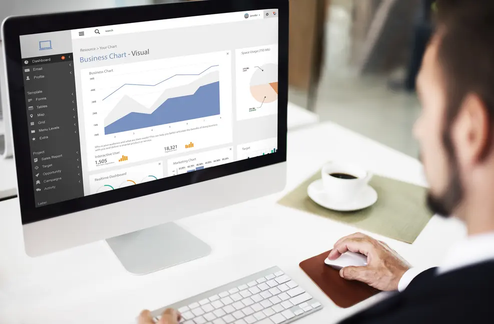 Mann, der an einem Computer mit einem übersichtlichen Dashboard arbeitet, das Diagramme und Statistiken anzeigt. Auf dem Tisch steht eine Tasse Kaffee, was eine produktive Arbeitsumgebung suggeriert. Das Bild vermittelt das Thema der Datenanalyse und digitalen Entscheidungsfindung im modernen Geschäftsbetrieb.