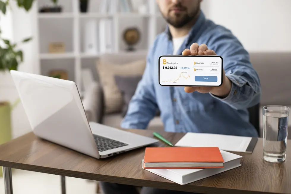 Mann zeigt auf seinem Smartphone eine Bitcoin-Wallet-App mit aktuellen Preisangaben, während er an einem Tisch mit einem Laptop, Notizblock und Glas Wasser sitzt. Hintergrund mit Pflanzen und Bücherregal, Atmosphäre der digitalen Finanzverwaltung und des Remote-Arbeitens.