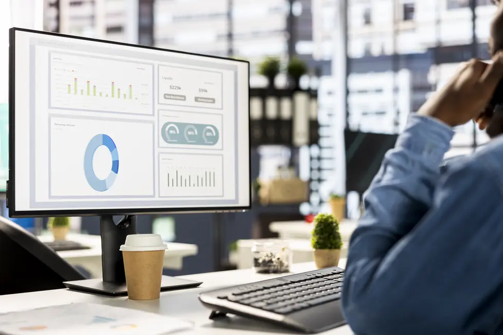 Mann sitzt an einem Arbeitsplatz und analysiert Geschäftsdaten auf einem Monitor. Der Bildschirm zeigt verschiedene Diagramme und Statistiken. Neben der Tastatur steht ein Pappbecher mit Deckel. Im Hintergrund sind Pflanzen und Büroartikel sichtbar, die eine produktive Arbeitsumgebung schaffen.