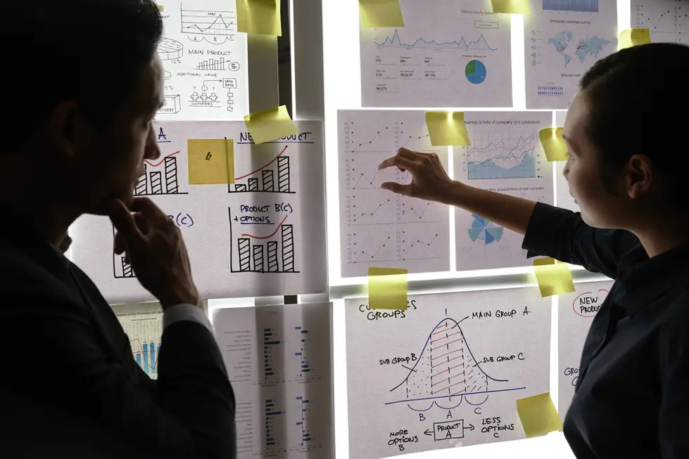 Zwei Personen betrachten aufmerksam ein Poster mit verschiedenen Diagrammen und Grafiken, während sie gemeinsam strategische Schritte zur Implementierung von strukturierten Daten erarbeiten. Die Präsentation zeigt verschiedene Analyseergebnisse, darunter Balkendiagramme, Liniendiagramme und statistische Informationen. Auf einigen Blättern befinden sich gelbe Haftnotizen, die wichtige Punkte hervorheben. Diese Szene verdeutlicht den kollaborativen Prozess der Datenanalyse und -planung für eine erfolgreiche Umsetzung von Schema Markup in einem Unternehmenskontext.