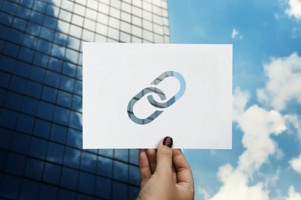 Hand hält ein Blatt Papier mit einem stilisierten Linksymbol vor einem modernen, reflektierenden Gebäudefenster und einem blauen Himmel. Das Bild vermittelt das Thema Linkaufbau und Digitalisierung.