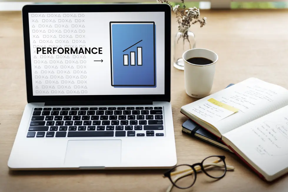 Laptop mit einem Bildschirm, der die Wortmarke "PERFORMANCE" und ein Diagramm zeigt, das auf eine Steigerung hinweist. Neben dem Laptop befinden sich eine Tasse Kaffee, ein Notizbuch mit handgeschriebenen Notizen und eine Brille. Der Hintergrund zeigt eine holzfarbene Oberfläche, die warm und einladend wirkt.