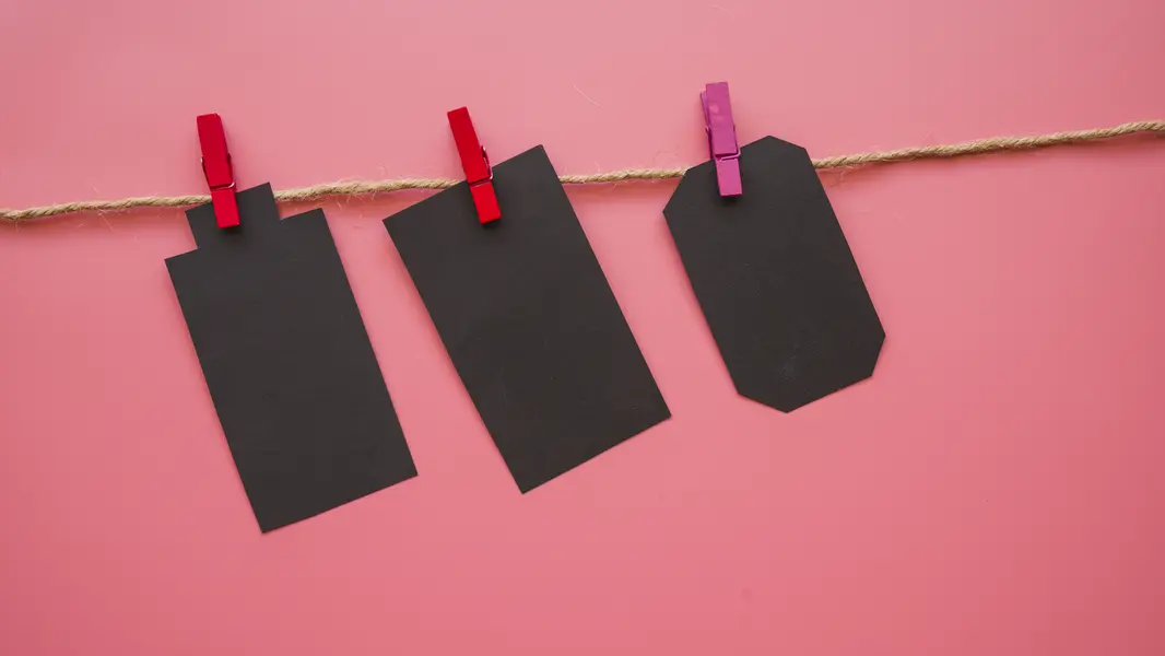 Drei schwarze Etiketten hängen an einer Juteschnur, die mit roten und rosa Wäscheklammern befestigt sind. Der Hintergrund ist in einem sanften Pink gehalten. Diese Anordnung eignet sich ideal für kreative DIY-Projekte, Preisgestaltung oder zur Personalisierung von Geschenken. Die Verwendung von verschiedenen Formen und Farben der Klammern verleiht dem Bild einen lebhaften und ansprechenden Charakter.