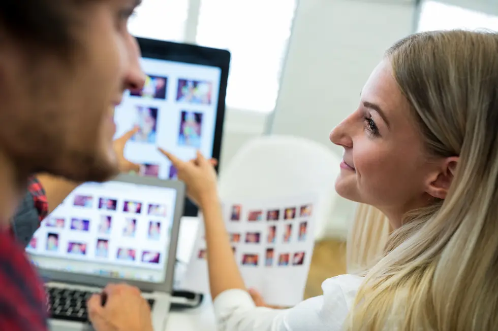 Zwei Personen, eine Frau mit langen blonden Haaren und ein Mann, interagieren in einem kreativen Arbeitsumfeld. Die Frau lächelt und schaut den Mann an, während sie auf ein Blatt Papier zeigt, das Fotos enthält. Auf einem Laptop im Vordergrund sind ebenfalls Bildvorschauen zu sehen. Die Szene vermittelt ein Gefühl von Zusammenarbeit und Kreativität im Designprozess.
