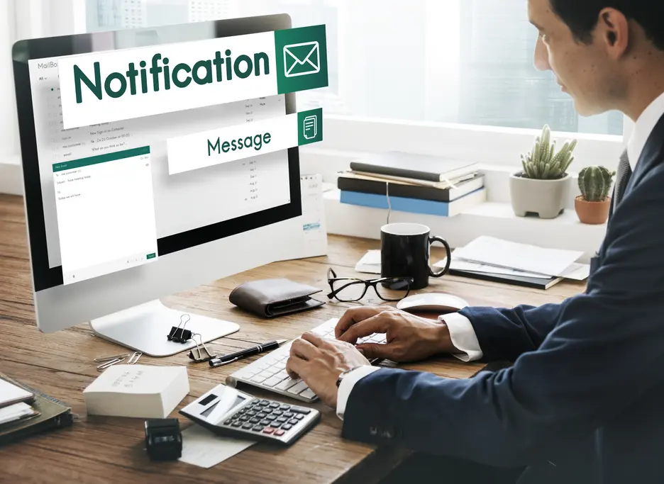 Mann im Anzug sitzt an einem Schreibtisch und tippt an einem Computer. Auf dem Bildschirm ist eine E-Mail-Benachrichtigung zu sehen, die den Text „Notification“ und „Message“ zeigt. Der Schreibtisch ist mit Büromaterialien, einem Kaffeebecher und einer Pflanze dekoriert. Sichtbar sind auch ein Taschenrechner und Notizen auf dem Tisch.
