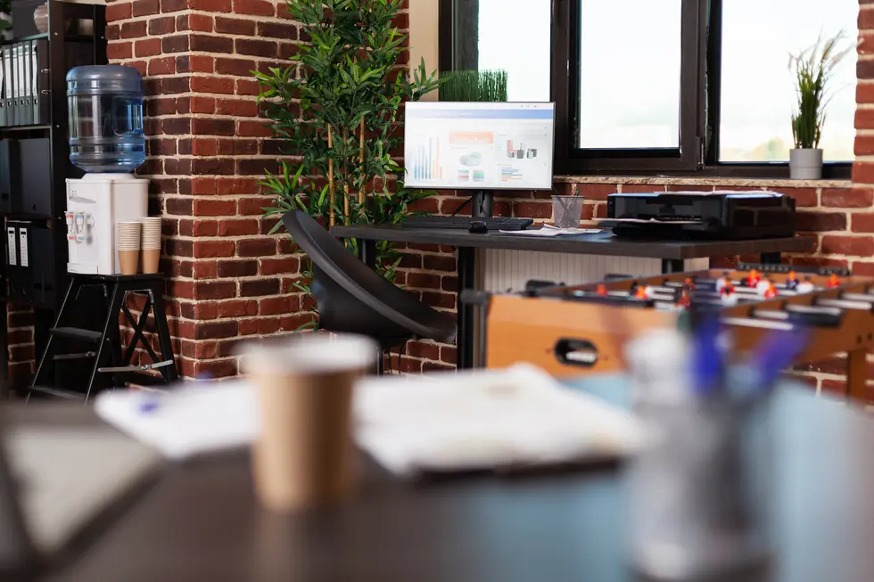 Moderner Arbeitsplatz mit einem Schreibtisch, an dem ein Computer mit Analysediagrammen sichtbar ist. Im Hintergrund befindet sich ein Wasserspender sowie eine Pflanze, während auf dem Tisch Papiere und ein Kaffeebecher zu sehen sind. Die Backsteinwand sorgt für eine angenehme Atmosphäre im Büro.