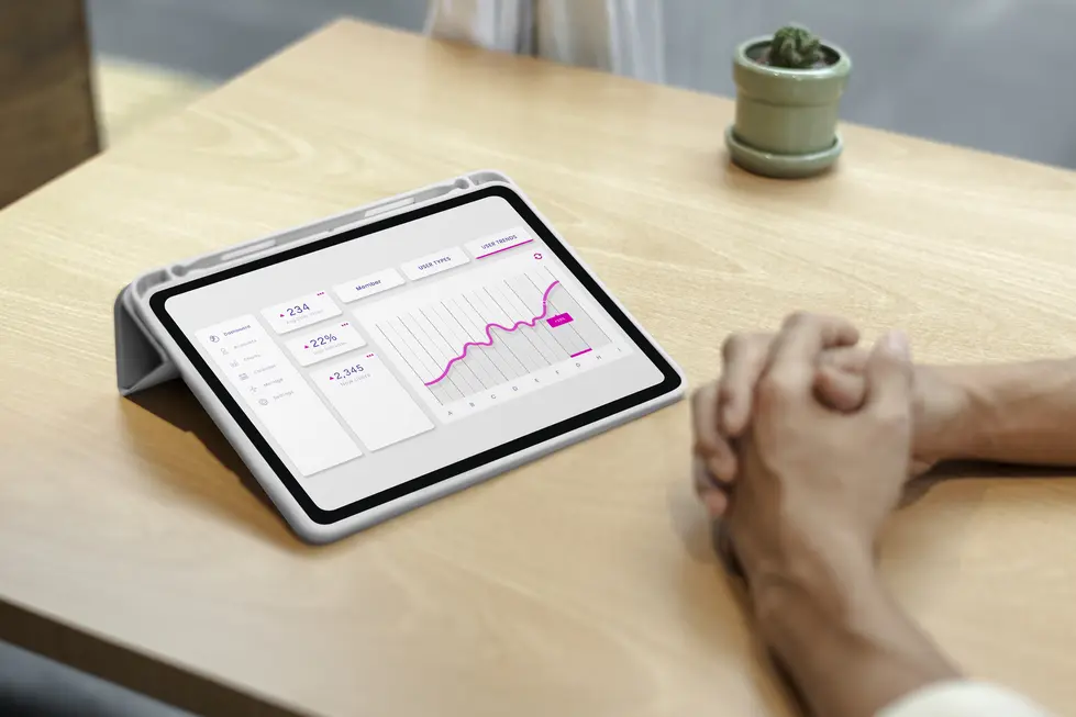 Tablet auf Holzoberfläche zeigt ein Analyse-Dashboard mit grafischen Darstellungen von Nutzerstatistiken, darunter Nutzersaldo, Wachstumsprozent und neue Nutzer. Die Benutzeroberfläche ist in einem modernen, minimalistischen Design gehalten, mit einer grafischen Darstellung in pinken und grauen Tönen. Hände einer Person sind im Vordergrund zu sehen, während ein kleiner Blumentopf im Hintergrund platziert ist. Ideal für die Illustration von Datenanalyse und Web-Traffic-Überwachung.