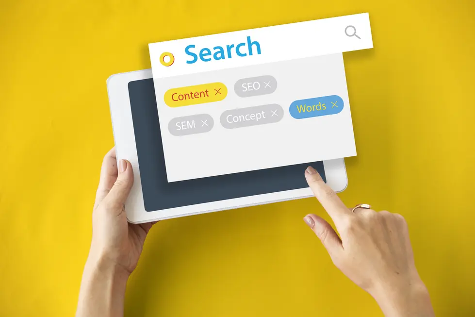 Hände halten ein Tablet, auf dem eine Suchmaske angezeigt wird. Der Bildschirm zeigt Begriffe wie "Content", "SEO", "SEM", "Concept" und "Words", wobei einige davon markiert sind. Der Hintergrund ist gelb, was das Bild hell und einladend wirken lässt.
