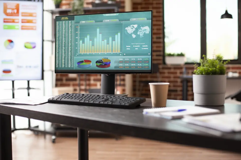 Moderne Arbeitsplatzumgebung mit Computerbildschirm, der umfassende Datenanalysen und Diagramme zeigt. Der Schreibtisch ist ordentlich mit einem Keyboard, einem Notizblock und einem Becher Kaffee dekoriert. Im Hintergrund ist ein zweiter Bildschirm sichtbar, der weitere Datenvisualisierungen anzeigt. Die Szenen sind in einem warmen, großzügigen Raum mit Fensterbelichtung und Pflanzen gestaltet, die eine frische und anregende Atmosphäre schaffen. Ideal für datengetriebenes Arbeiten und Team-Kollaboration.