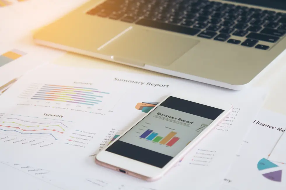 Smartphone zeigt einen Business Report auf einem Tisch mit gedruckten Zusammenfassungs- und Finanzberichten sowie einem Laptop im Hintergrund. Die Berichte enthalten Diagramme und Statistiken zur Analyse von Verkaufsdaten und Kunden.