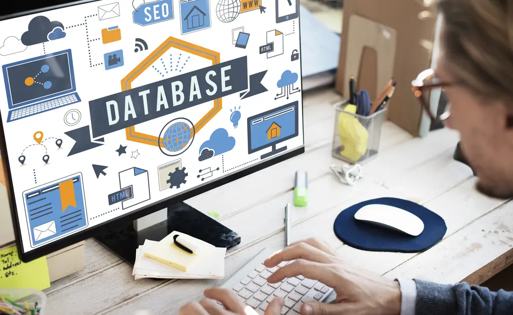Mann, der an einem Schreibtisch arbeitet und auf einen Computerbildschirm blickt, auf dem das Wort "DATABASE" in großen Buchstaben zu sehen ist. Um das Wort herum sind verschiedene Symbole und Icons, die mit SEO, HTML und Datenbanken in Verbindung stehen. Der Schreibtisch ist mit unterschiedlichen Büromaterialien wie Stiften, Notizen und einem Computermauspad ausgestattet. Die Szene vermittelt ein modernes Arbeitsumfeld im Bereich Webentwicklung oder digitales Marketing.