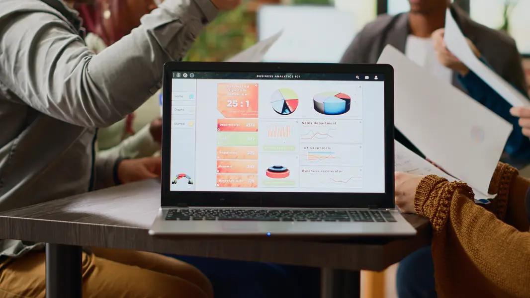 Team von Fachleuten arbeitet zusammen an einem Laptop, der eine benutzerfreundliche Business-Analytics-Oberfläche zeigt, auf der wichtige Kennzahlen wie Umsatzprognosen, Vertriebsergebnisse und analytische Diagramme visualisiert sind. Im Hintergrund sind diskutierende Personen zu sehen, mientras sie Unterlagen in den Händen halten.