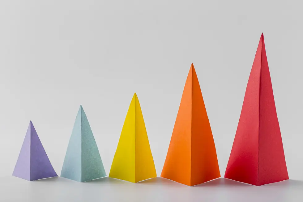 Bunt gefaltete Papierpyramiden in verschiedenen Größen und Farben stehen nebeneinander auf einem hellen Hintergrund. Die Farbtöne reichen von Lila über Blau und Gelb bis zu Orange und Rot, was eine lebendige Darstellung von Vielfalt und Kreativität zeigt.