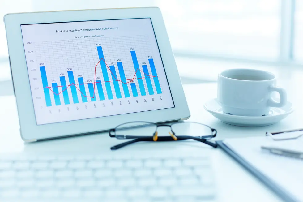 Dashboard mit Geschäftsdaten auf einem Tablet, das Balkendiagramme und eine rote Trendlinie zur Anzeige der betrieblichen Aktivitäten zeigt; im Hintergrund eine Tasse Kaffee und eine Tastatur, die eine moderne Büroumgebung symbolisieren.
