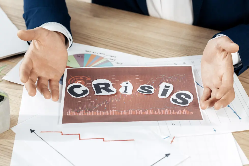 Hand eines Mannes, die auf einem Tisch mit mehreren Dokumenten und Grafiken liegt, während sie ein Blatt mit dem Wort "CRISIS" in großen Buchstaben präsentiert. Im Hintergrund sind Diagramme und analytische Daten sichtbar, die auf eine Krisensituation hinweisen, was auf ein Meeting zur Krisenbewältigung deuten könnte.