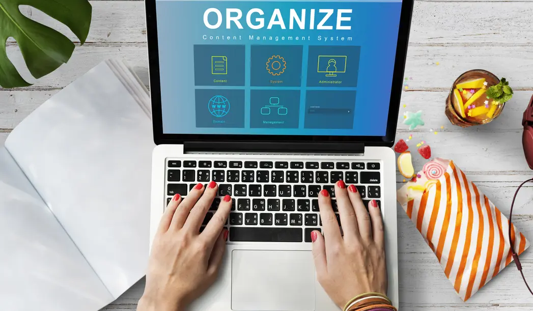 Hände tippen auf einer Laptop-Tastatur, während auf dem Bildschirm eine Benutzeroberfläche für ein Content Management System mit der Aufschrift "ORGANIZE" und verschiedenen Optionen wie "Content", "System", "Administrator", "Domain" und "Management" angezeigt wird. Neben dem Laptop liegen eine offene Zeitschrift und eine Tüte mit bunten Süßigkeiten, während ein Getränk mit Fruchtstücken neben dem Laptop steht.