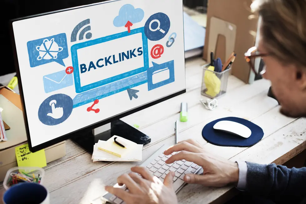 Mann tippt an einer weißen Tastatur vor einem Monitor, auf dem das Wort "BACKLINKS" in blauer Schrift zu sehen ist. Um das Wort herum sind verschiedene Symbole, die mit digitalem Marketing und Linkbuilding in Verbindung stehen, darunter Wolken, Suchsymbol und Klick-Icons. Der Arbeitsplatz ist unordentlich, mit Büromaterial und Post-its auf dem Tisch.