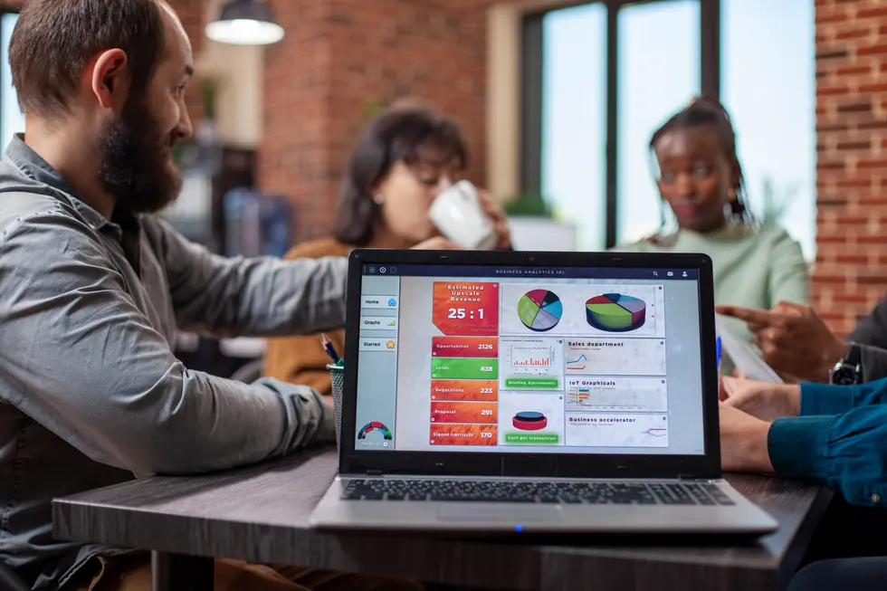 Laptop mit einer Business-Analytics-Oberfläche, die Umsatzprognosen und Diagramme zeigt, während drei Personen an einem Tisch diskutieren, zwei davon sind konzentriert bei einer Präsentation, während eine Person eine Tasse hält und im Hintergrund sichtbar ist, mit einer gemütlichen Büroumgebung.