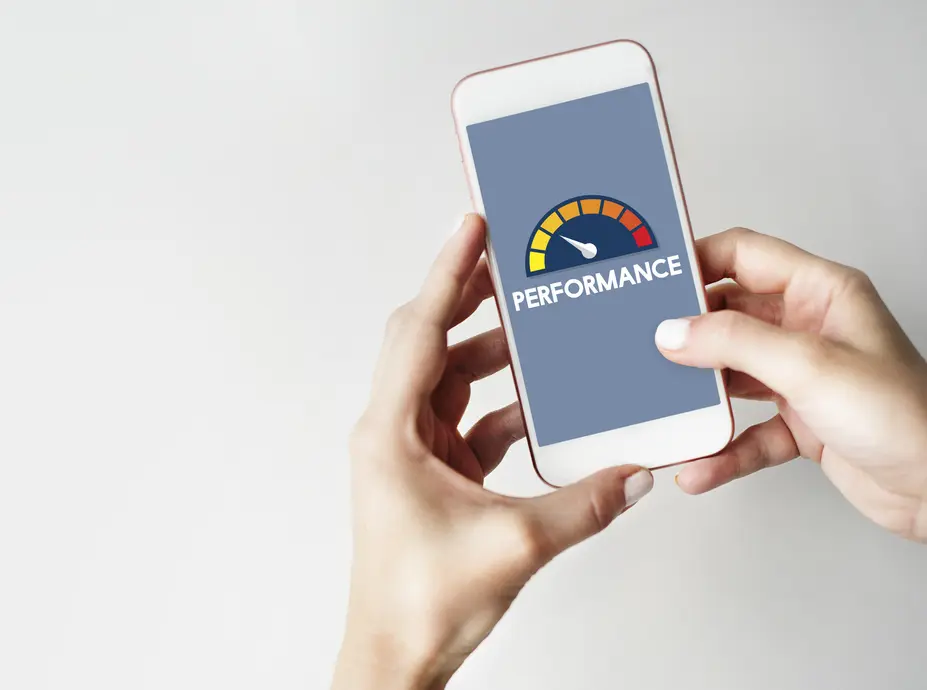 Eine Person hält ein Smartphone in der Hand, auf dessen Bildschirm ein Performance-Diagramm mit der Beschriftung "PERFORMANCE" zu sehen ist. Das Diagramm zeigt einen Indikator, der von grün über gelb bis rot reicht, um die Leistung zu visualisieren. Der Hintergrund des Bildschirms ist grau, während das Smartphone auf einer hellen Oberfläche positioniert ist.