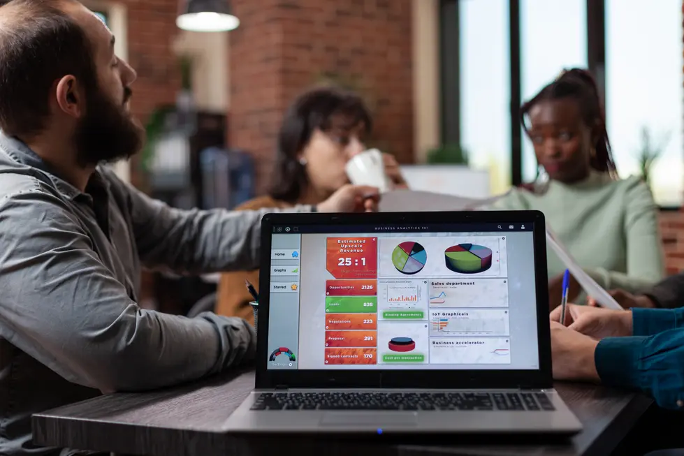 Gruppensitzung in einem modernen Büro, bei der ein Team an einem Laptop arbeitet, der Datenanalysen und Diagramme zeigt. Auf dem Bildschirm sind wichtige Metriken wie Umsatzprognosen, Anzahl der Möglichkeiten und Diagramme zu sehen, während im Hintergrund Teammitglieder diskutieren und Notizen machen. Der Fokus liegt auf der strategischen Nutzung von Daten für geschäftliche Entscheidungen.