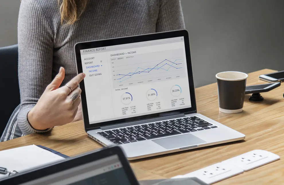Eine Person zeigt mit dem Finger auf ein Laptop-Display, das ein Finance-Dashboard anzeigt. Das Dashboard umfasst Diagramme zur Einkommens- und Ausgabenanalyse sowie statistische Informationen zu Gesamtumsätzen. Auf dem Tisch befinden sich ein Kaffeebecher, ein Smartphone und Dokumente.