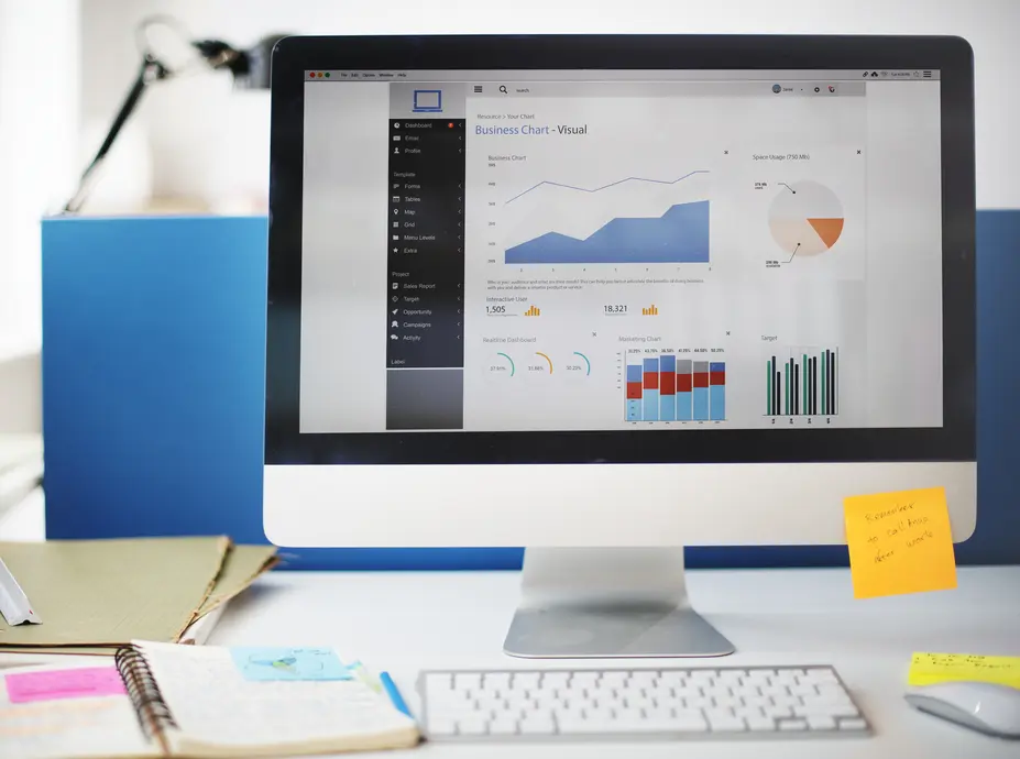 Ein moderner Computer-Bildschirm zeigt ein detailliertes Dashboard mit verschiedenen Analyse- und Grafikdiagrammen für Geschäftskennzahlen. Auf dem Bildschirm sind Verlaufskurven, Balkendiagramme und wichtige Statistiken wie interaktive Benutzer und Raumnutzung abgebildet. Neben dem Computer liegt ein Notizblock mit bunten Haftnotizen, der zusätzlich Informationen enthält.