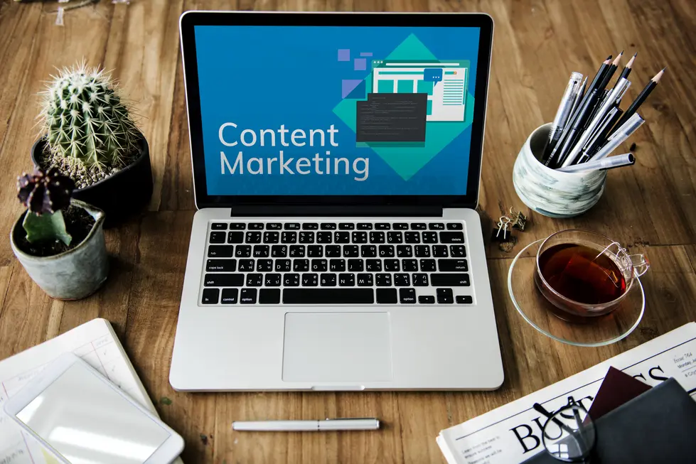 Laptop mit dem Bildschirmtext "Content Marketing" auf einem Holztisch, umgeben von einem Kaktus im Topf, Schreibutensilien in einem Behälter, einer Tasse Tee auf einem Untersetzer, einem Smartphone und offenen Notizen für Planungen.